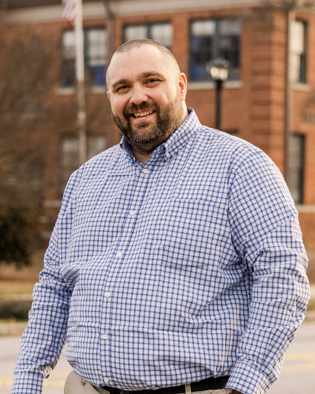 Kerry Hatcher, smiling, in a checkered shirt, standing on a curb in front of a brick public school building.